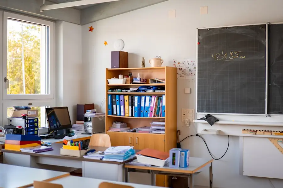Leeres Klassenzimmer mit Tafel, Aktenordnern im Regal und Heften auf den Tischen