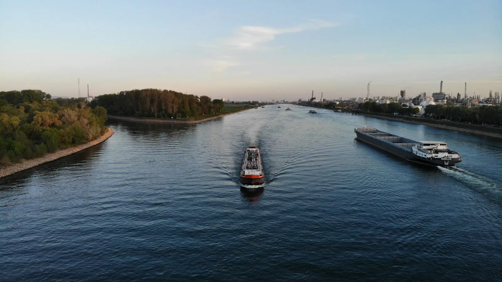 Luftaufnahme zweier Binnenschiffe auf einem Fluss zwischen bewaldetem Ufer und Industrieanlagen