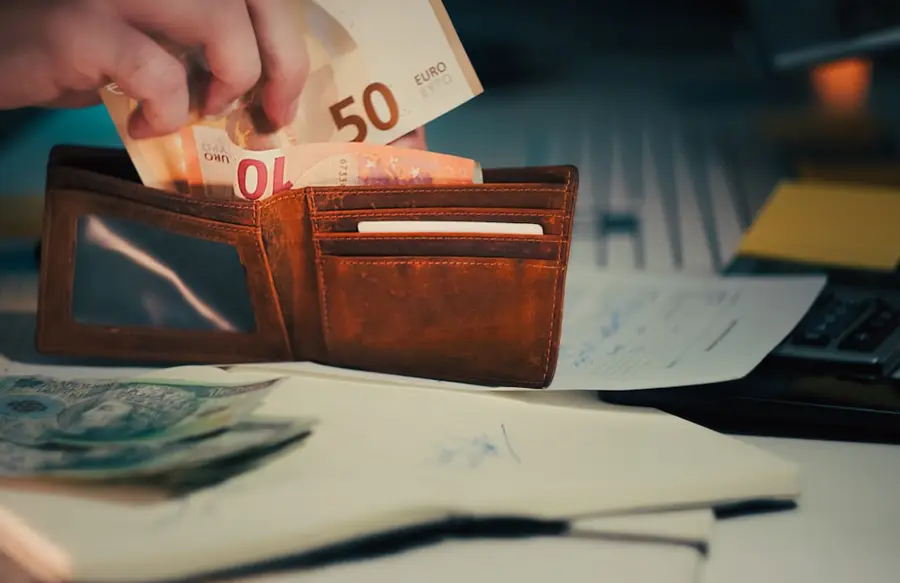 Hand steckt Euroscheine in ein Portemonnaie, daneben Unterlagen und ein Taschenrechner