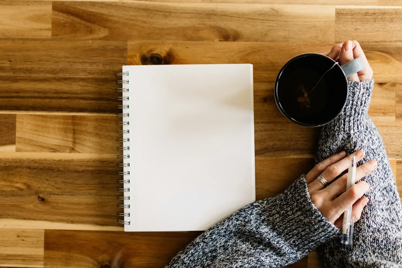 Ein Notizbuch liegt auf einem Holztisch; eine Person hält einen Stift und eine Tasse.