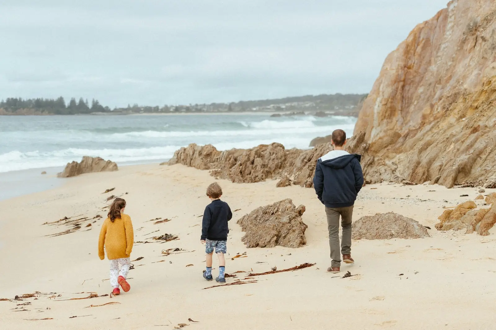 Ein Erwachsener und zwei Kinder gehen am Meer entlang, von hinten gesehen