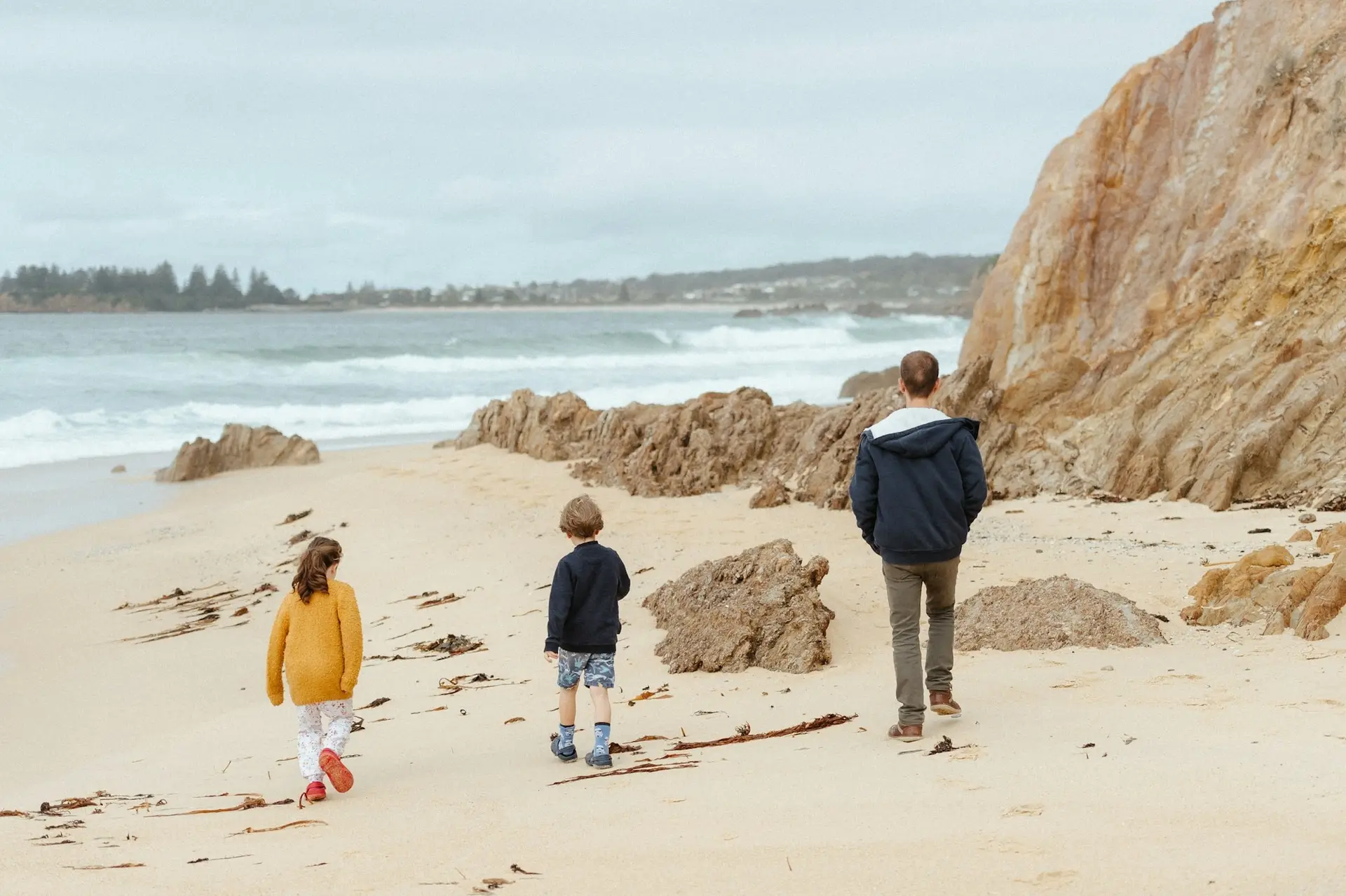 Ein Erwachsener und zwei Kinder gehen am Meer entlang, von hinten gesehen