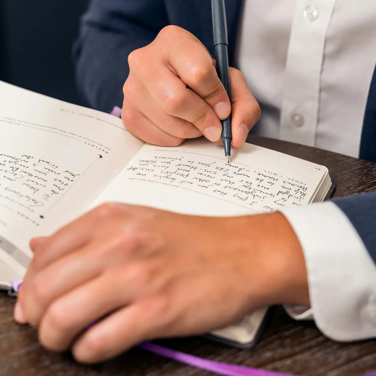 Hand hält einen Stift und schreibt in ein aufgeschlagenes Notizbuch, daneben liegt die zweite Hand auf dem Tisch